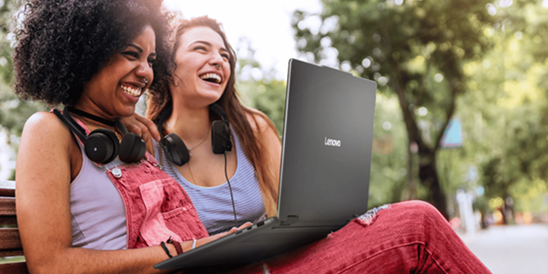 Two women on a park bench wearing headphones and browsing on a Lenovo ThinkPad
